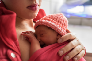 Small infant kangarooing on their mothers chest, wearing a red knitted hat and secure under a blanket. The mother has put her hand in the baby's back.